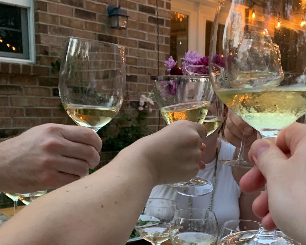 A toast: hands raising glasses of white wine at an outdoor table under string lights